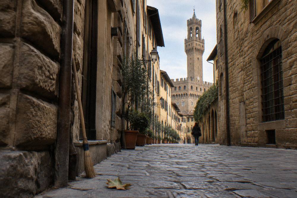 Looking up the crenellated stone tower of Palazzo Vecchio from the narrow Via de' Gondi, high rusticated walls on one side, a fallen plane leaf on the cobbles, a leaning broom