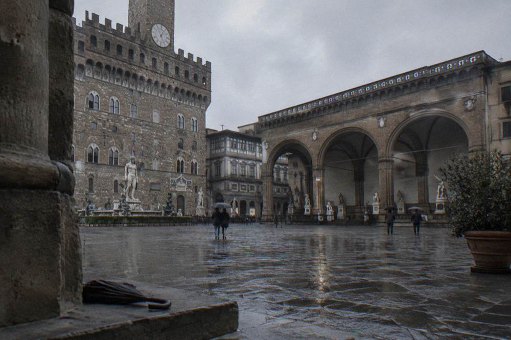 The open stone arches of the Loggia dei Lanzi beside Palazzo Vecchio seen across the wet square, small dark shapes inside the arches, wet paving reflecting, a folded umbrella on the step