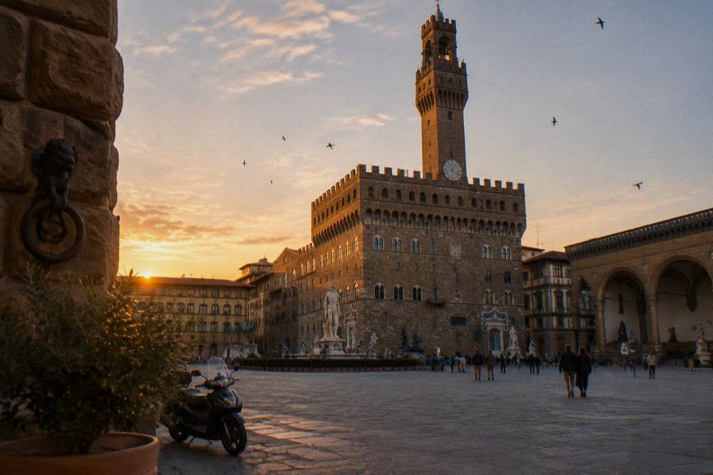 The rusticated pietra forte facade of Palazzo Vecchio with its crenellated tower rising above terracotta rooftop lines, warm low sun catching the stone, swifts wheeling