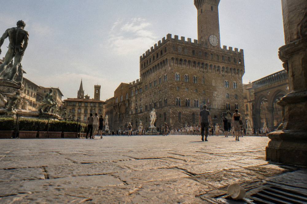 Low across the worn trachyte paving of Piazza della Signoria, the rusticated stone walls and crenellated tower of Palazzo Vecchio looming above, statues in open air