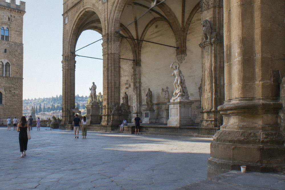 Seen from the far side of the square toward the Loggia dei Lanzi, its wide pietra forte arches sheltering marble figures, a discarded paper cup by a column base