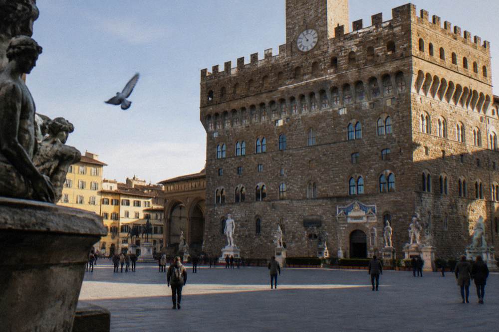 The rusticated facade of Palazzo Vecchio in raking light seen from across the square, statues on weathered plinths at street distance as small pale shapes, a pigeon drifting past