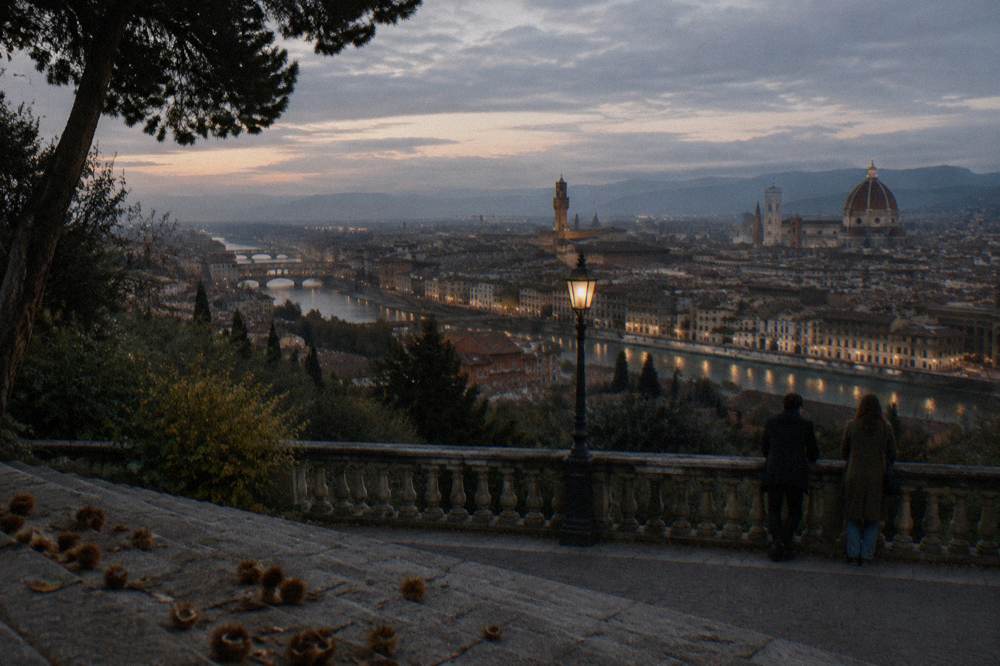 From behind two distant figures leaning on the Piazzale Michelangelo balustrade as the city's streetlamps flicker on below and the Arno turns pewter