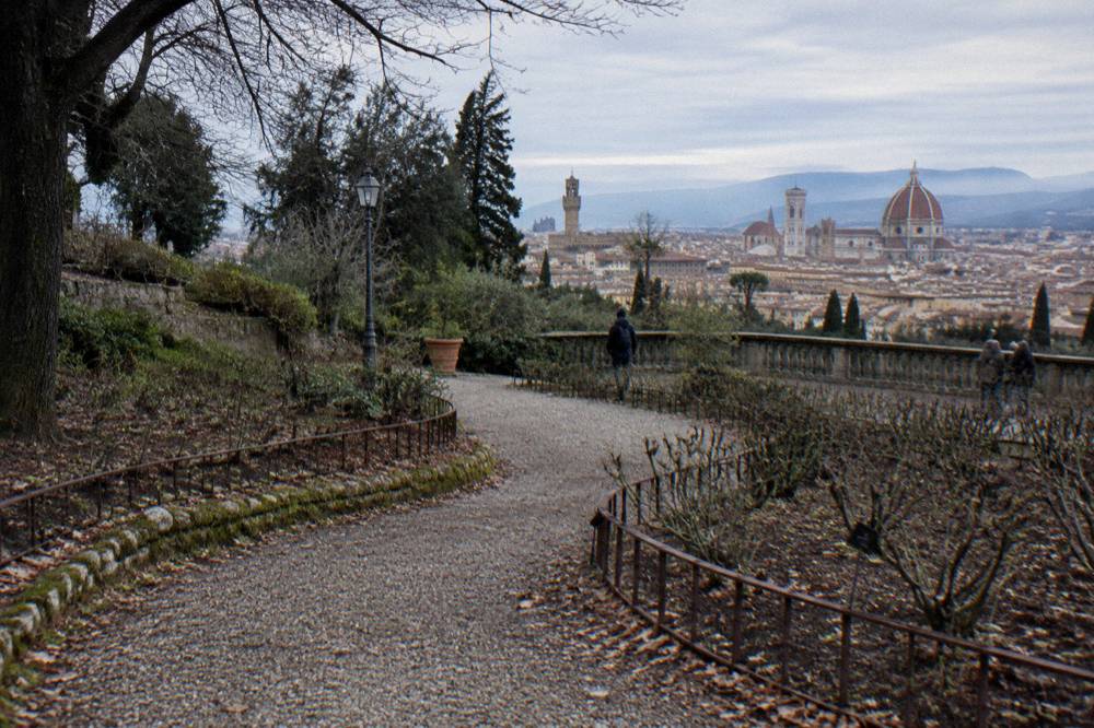 A gravel path curving down the hillside between bare rose beds edged with rusted iron, the Piazzale terrace balustrade visible above, dry leaves caught in the gravel