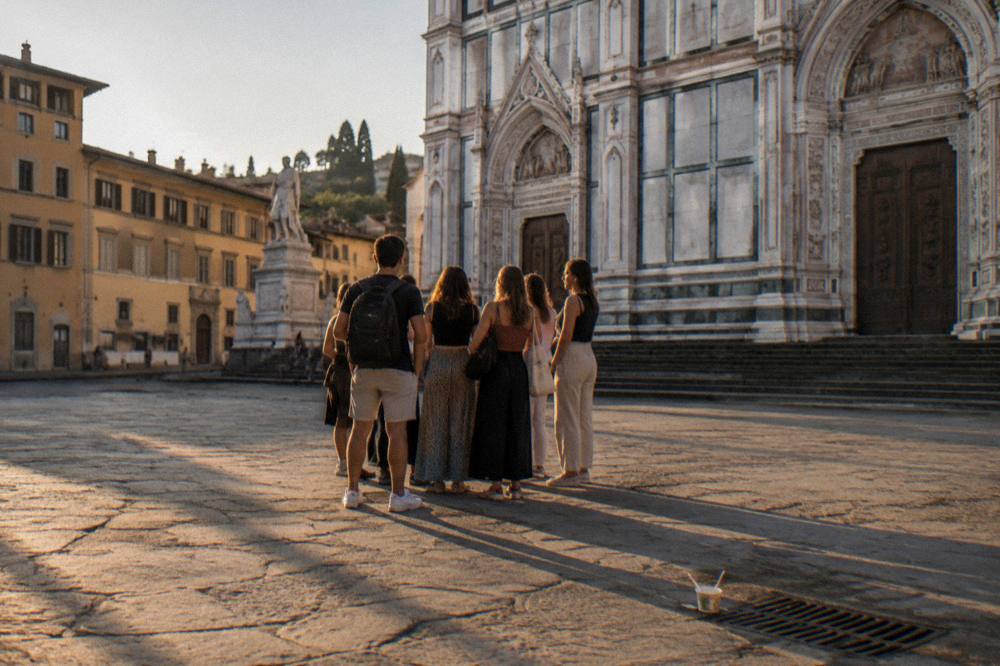 Warm September evening in Piazza Santa Croce, Florence, Italy, a small walking group gathered from behind near the church's striped marble facade, long shadows across worn stone paving