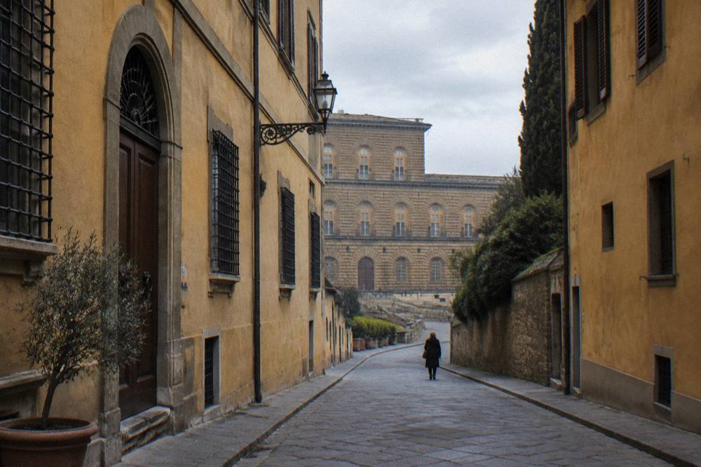 A narrow Via Guicciardini running toward the great rusticated block of Palazzo Pitti at its end, grey daylight, ochre plaster facades on both sides, one distant pedestrian from behind