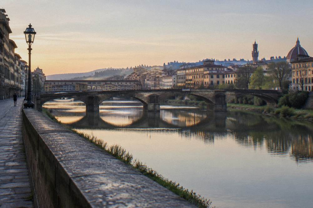 Pale Arno water reflecting the stone arches, the Ponte Vecchio downstream, dew on the parapet, a lone runner far off along the lungarno