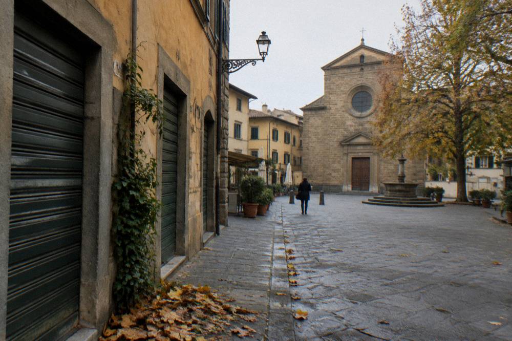Ochre plaster and pietra forte walls, closed green workshop shutters, fallen leaves gathered against a doorway, a distant figure with a coffee cup, Piazza Santo Spirito