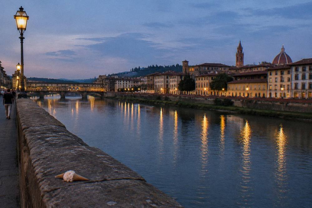 From Ponte alle Grazie looking along the water, lamplight smearing gold across the current, terracotta rooftops darkening, a discarded gelato cone on warm stone