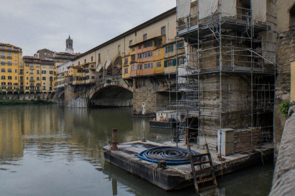 Low from the riverbank, scaffolding clamped to the stone piers beneath Ponte Vecchio, work pontoons moored on the slow Arno, coiled hoses and a rusted ladder on the nearest pontoon