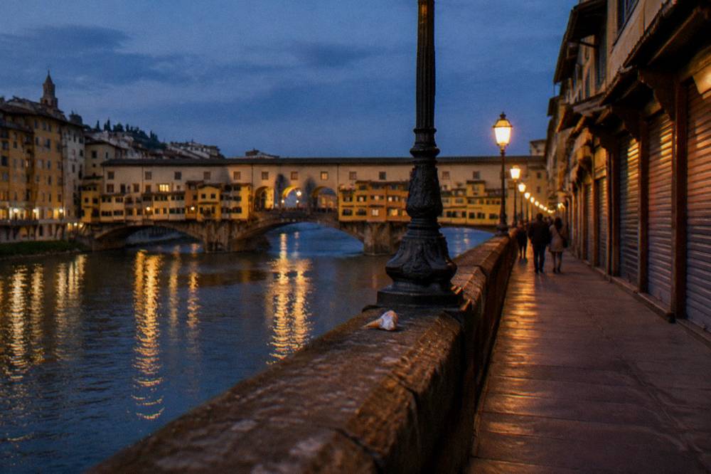 Looking along the parapet of Ponte Vecchio as its iron bridge lamps come on, gold light smearing across the dark Arno, closed shop shutters, a dropped ice-cream wrapper by a lamp base