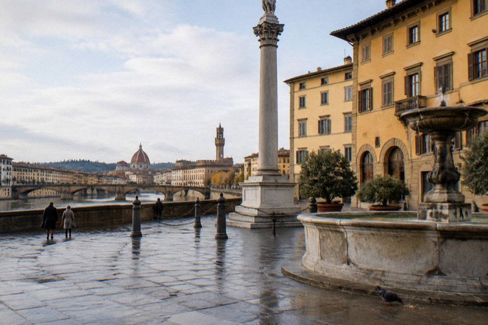 Across Piazza Santa Trinita toward its tall marble column and stone fountain, wet trachyte paving, a pigeon pecking at crumbs near the fountain basin, ochre palazzi behind