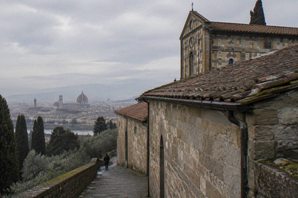 Soft grey April morning on the hill above Florence, Italy, low along the weathered stone facade and terracotta tiled roof of San Miniato abbey, damp masonry, moss in a gutter joint
