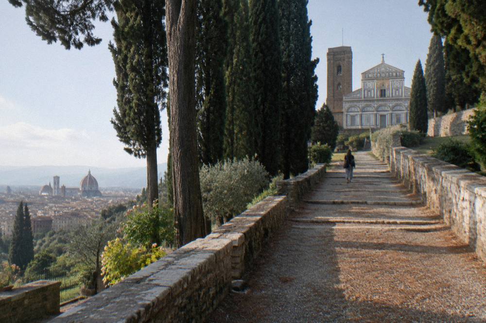 A gravel path climbing from the Rose Garden between dark cypress trees toward the abbey on the hill, pale stone walls flanking the steps, dry fallen needles scattered across the gravel