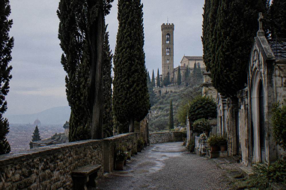 The tall stone bell tower of San Miniato rising behind the fortified hill wall, seen from the cemetery lane between dark cypress trees, grey sky, damp gravel, no one in frame