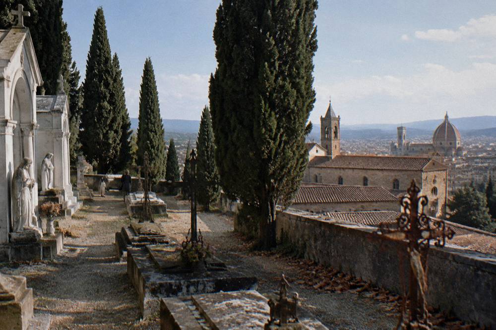 A walled cemetery terrace behind San Miniato al Monte, cypress trees standing among stone tombs and rusted iron crosses, terracotta roof of the abbey beyond