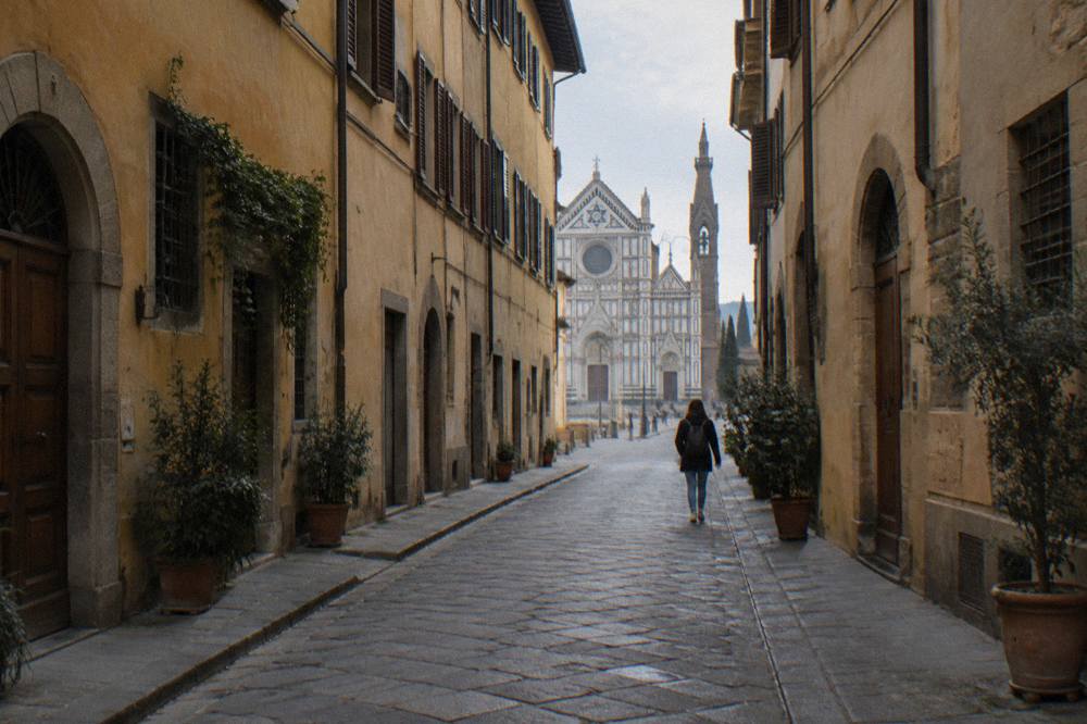 A narrow Borgo de' Greci street running toward the open end of Piazza Santa Croce, tall ochre palazzi on both sides, dusty light on the worn stone, a distant visitor from behind
