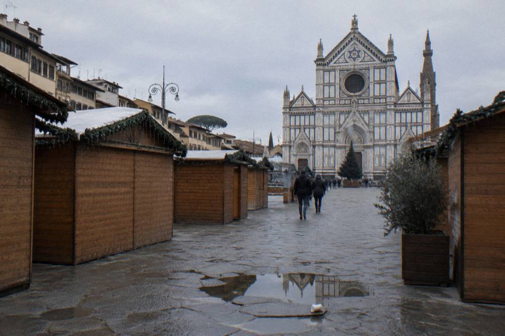 Wooden market huts lining the trachyte-paved square before the striped marble facade of Santa Croce, string lights unlit, a puddled patch of paving and a discarded paper cup