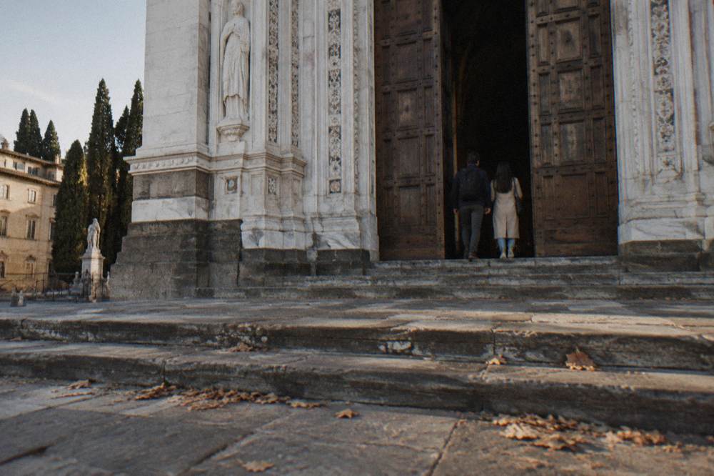 From low across the worn stone steps toward the tall wooden doors of Santa Croce basilica, two visitors from behind entering the shadowed doorway, dry leaves caught against the threshold