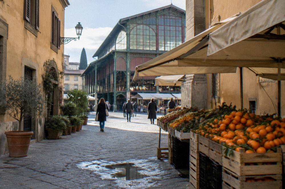 Looking down a narrow street toward the cast-iron market hall of Sant'Ambrogio, crates of oranges and canvas awnings outside ochre plastered facades, a puddle drying on the cobbles