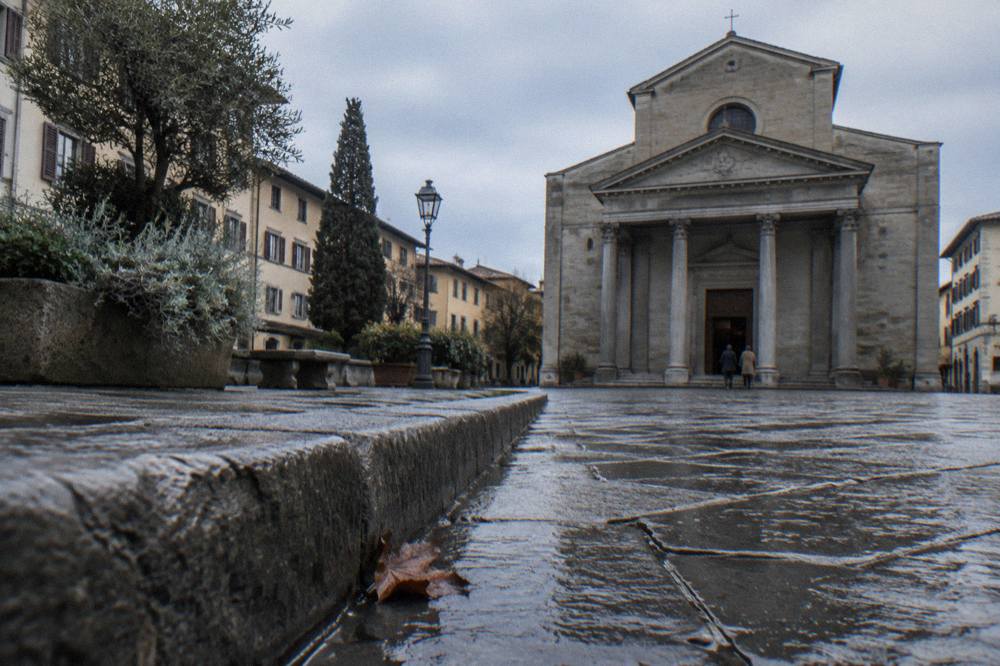 Low and close along the piazza's trachyte kerb toward the columned grey stone facade of Santo Spirito, two distant figures at the church door, a fallen leaf caught against the wet paving