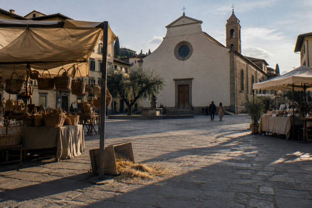 Low across the trachyte-paved square toward Santo Spirito's plain plastered facade, canvas craft stalls casting long shadows, a tipped wooden crate and scattered straw by a stall leg