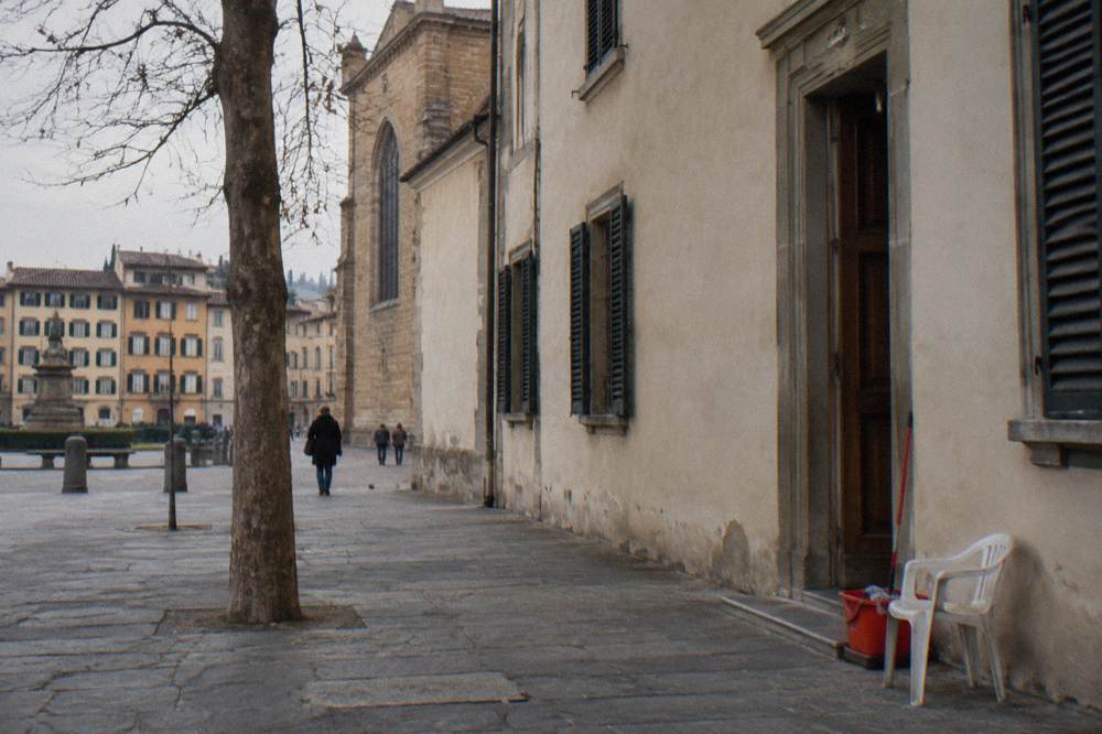 The plain plastered flank of the Santo Spirito church seen along a quiet side street, shuttered windows, a bare tree trunk, a plastic chair and a mop bucket left by a doorway
