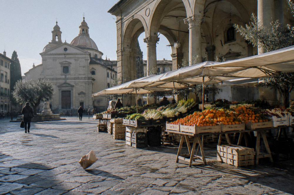 Low across the trachyte-paved square toward the open market stalls of the loggia, crates of clementines and chicory on trestles, a dropped paper bag rolling in the wind